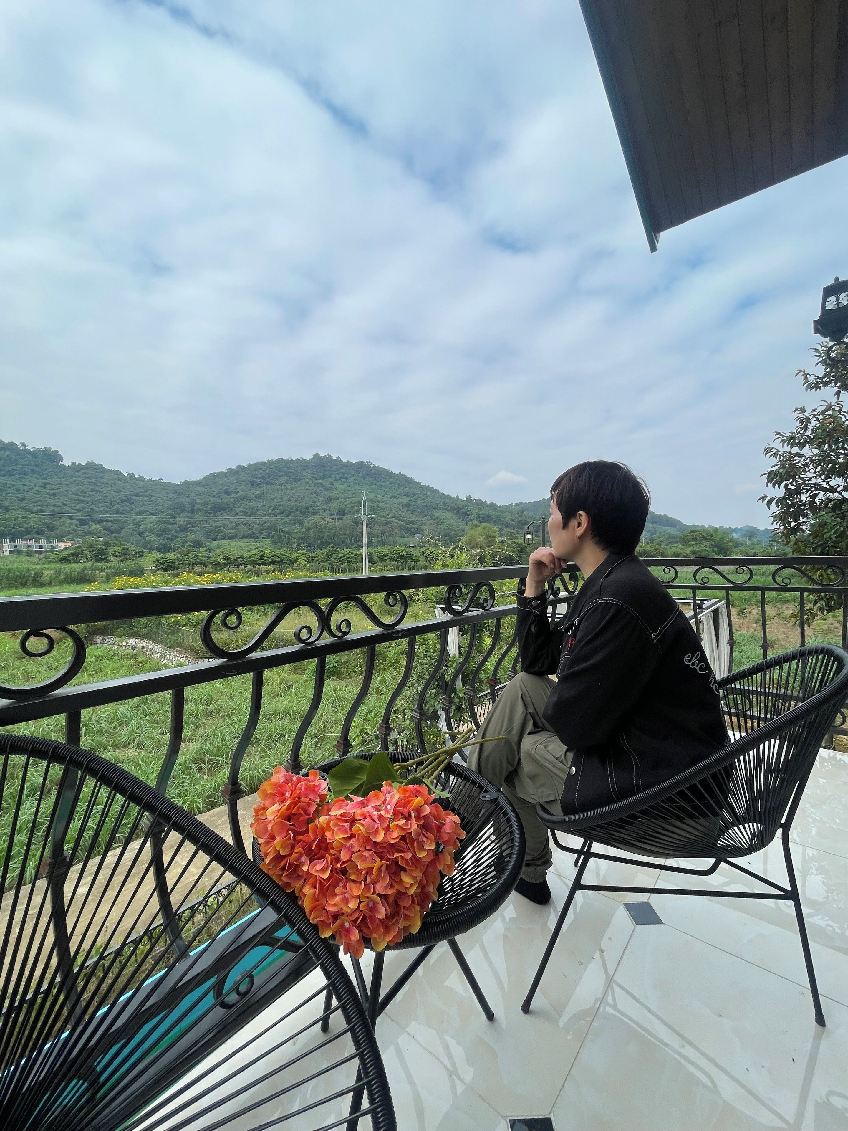 A person sitting on a black metal chair on a balcony with a view of green hills and a cloudy sky.
