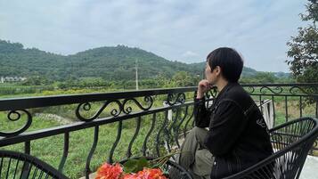 A person sitting on a black metal chair on a balcony with a view of green hills and a cloudy sky.