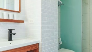 Un baño con pared de azulejos blancos, grifo negro, tocador de madera y cabina de ducha verde.