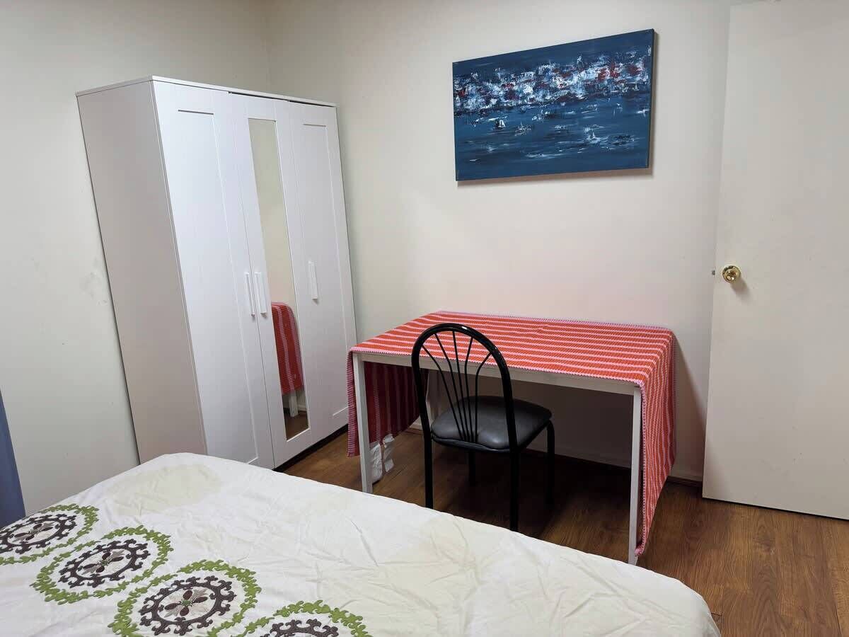 A bedroom with a bed, a desk with a chair, a wardrobe, and a painting on the wall.
