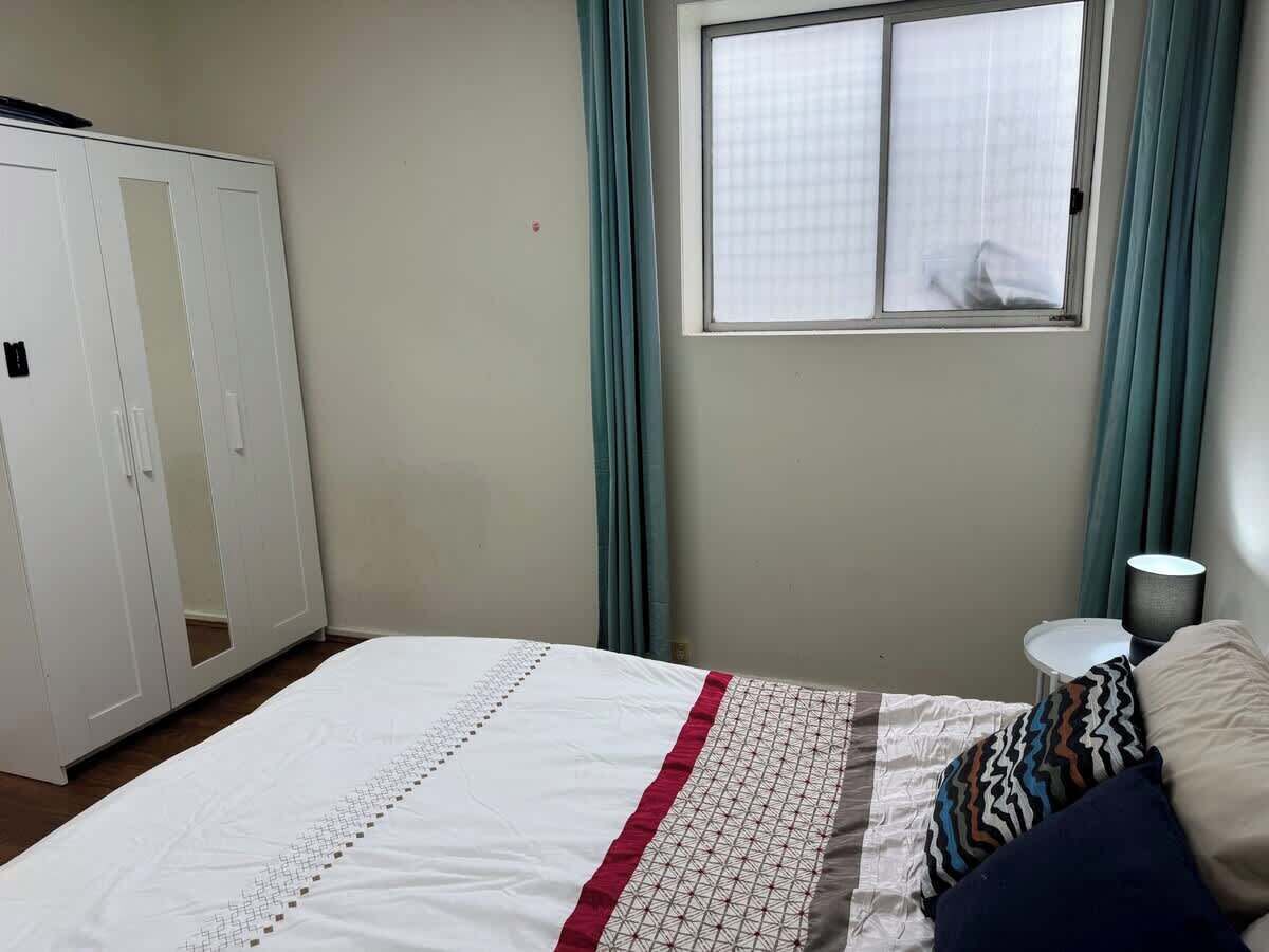 Een slaapkamer met een bed, een kledingkast, een raam met gordijnen en een nachtkastje met een lamp.