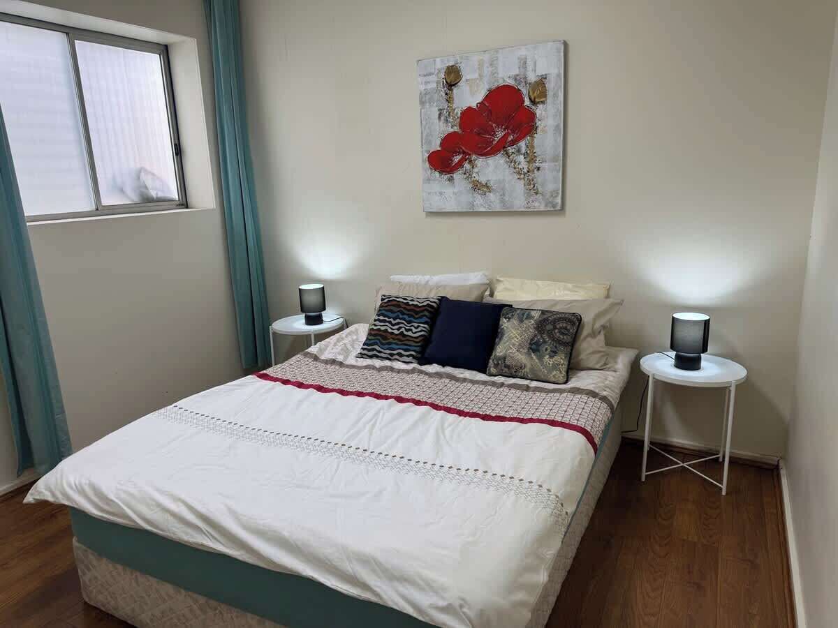 A neatly made bed with a white and teal bedspread, two bedside lamps, and a red abstract painting on the wall.