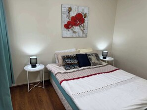 Een netjes opgemaakt bed met een wit-rode spreideken, met aan weerszijden bedlampen op witte nachtkastjes. Daarboven hangt een enkel kunstwerk met rode bloemen.