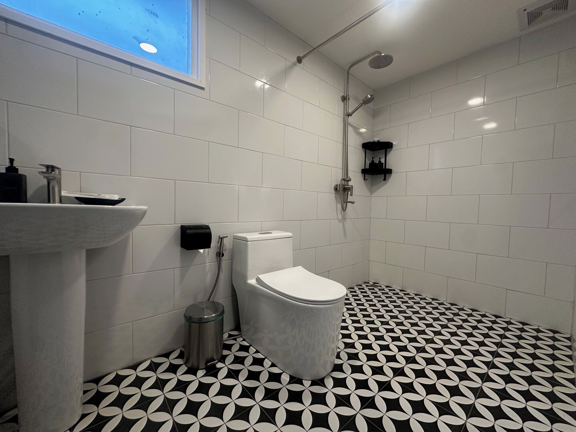 Un baño moderno con azulejos blancos, un suelo con estampado blanco y negro, un inodoro, un lavabo y una ducha.