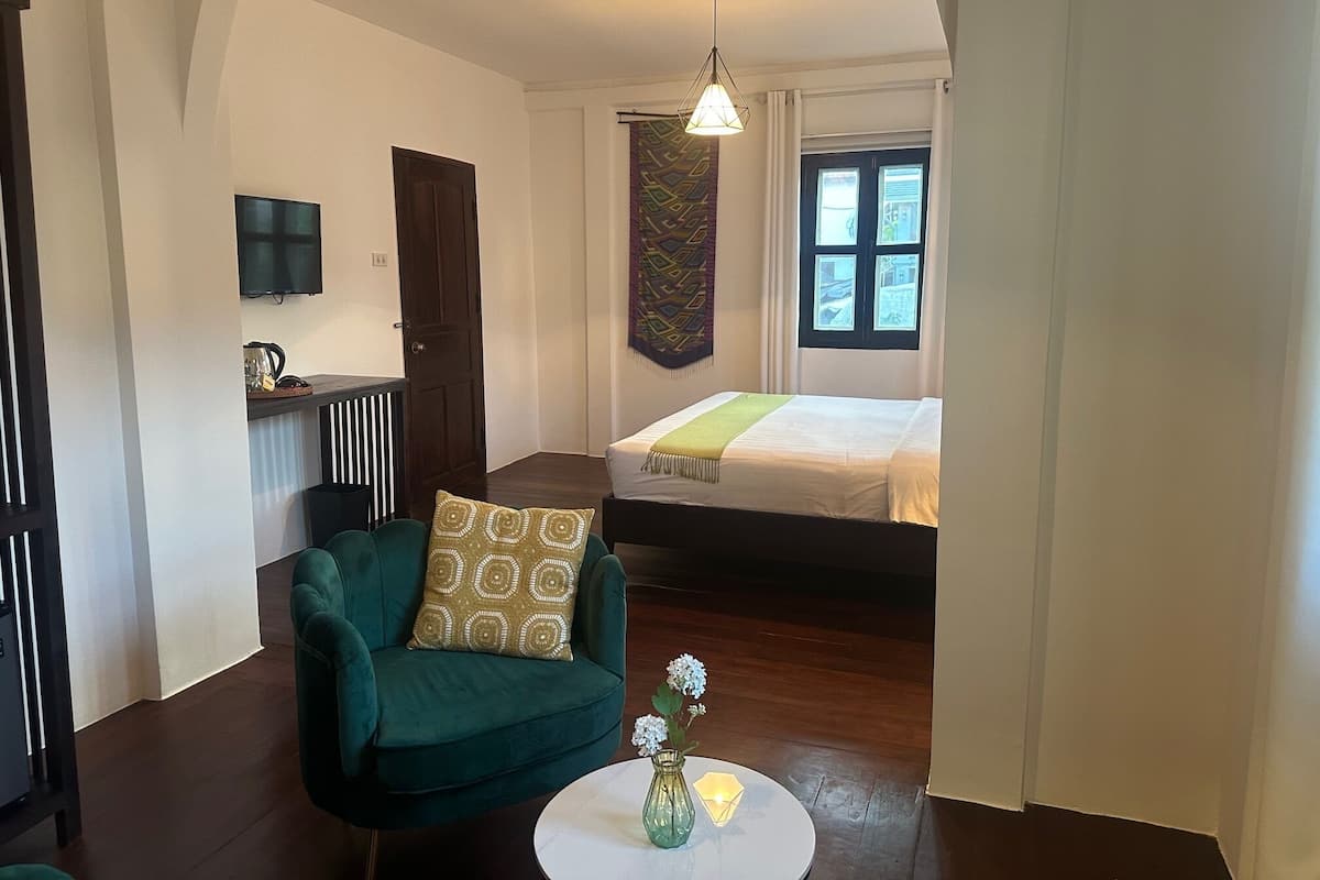 A compact hotel room with a green armchair, a small round table with a vase, a bed with a green blanket, a TV on the wall, and a window with curtains.