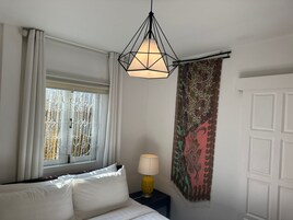 Un dormitorio con cama, ventana con cortinas, lámpara colgante, tapiz estampado y una puerta.