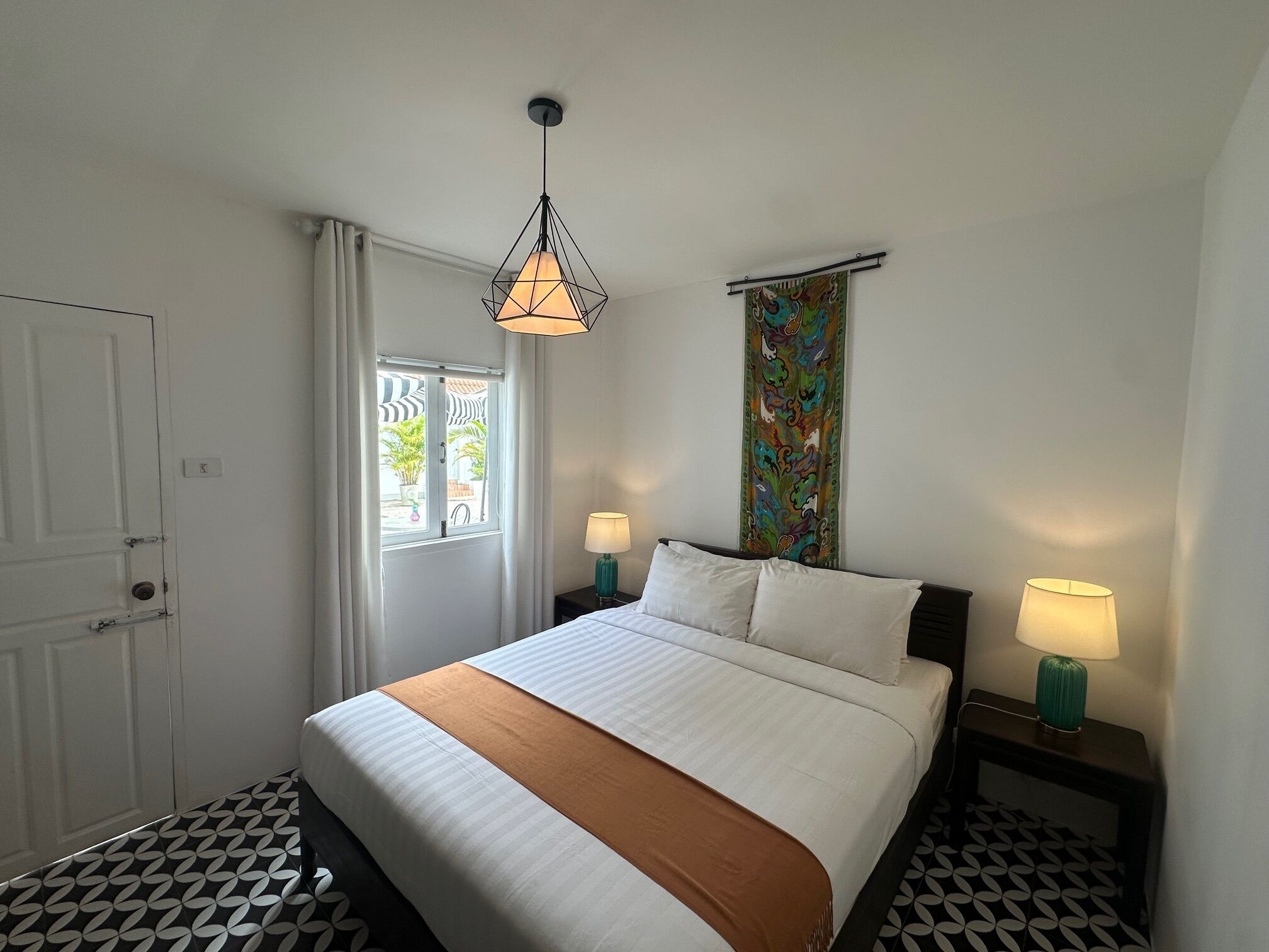 A hotel room with a bed, two bedside tables with lamps, a window with curtains, a door, and a patterned floor.