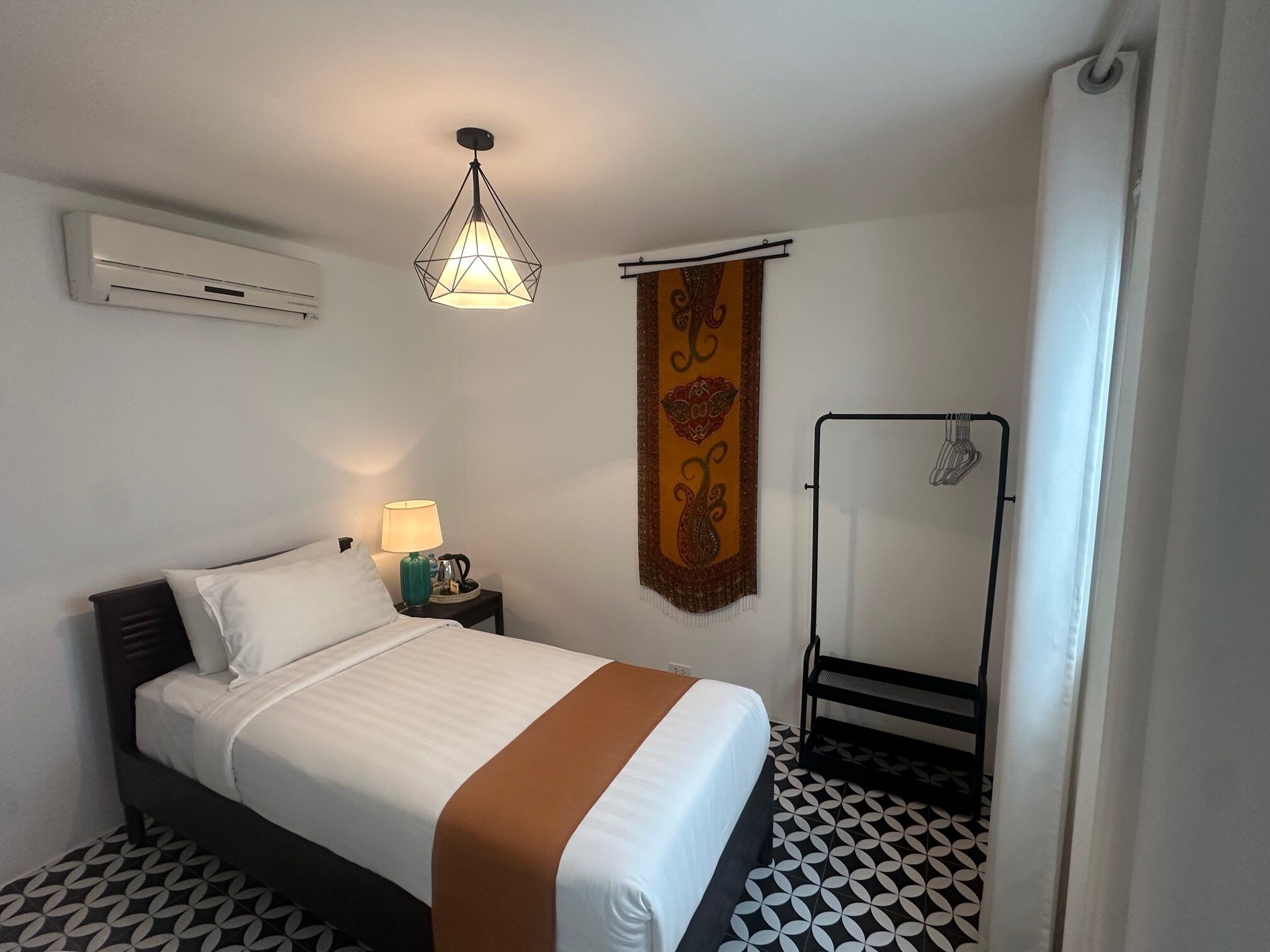 A hotel room with a bed, a nightstand, a hanging light fixture, a shower area, and a patterned floor.