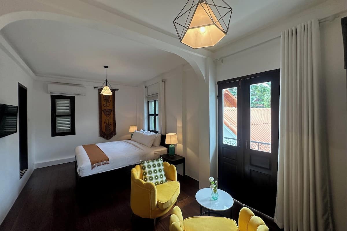 A hotel room with a bed, two yellow armchairs, a small table with a vase, a hanging light fixture, and a view of the outside through a door.