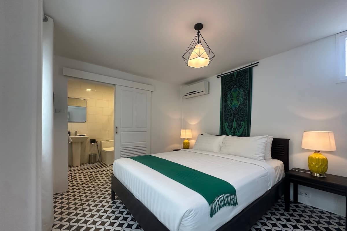 A hotel room with a bed, two bedside lamps, a green tapestry, and a geometric-patterned floor.