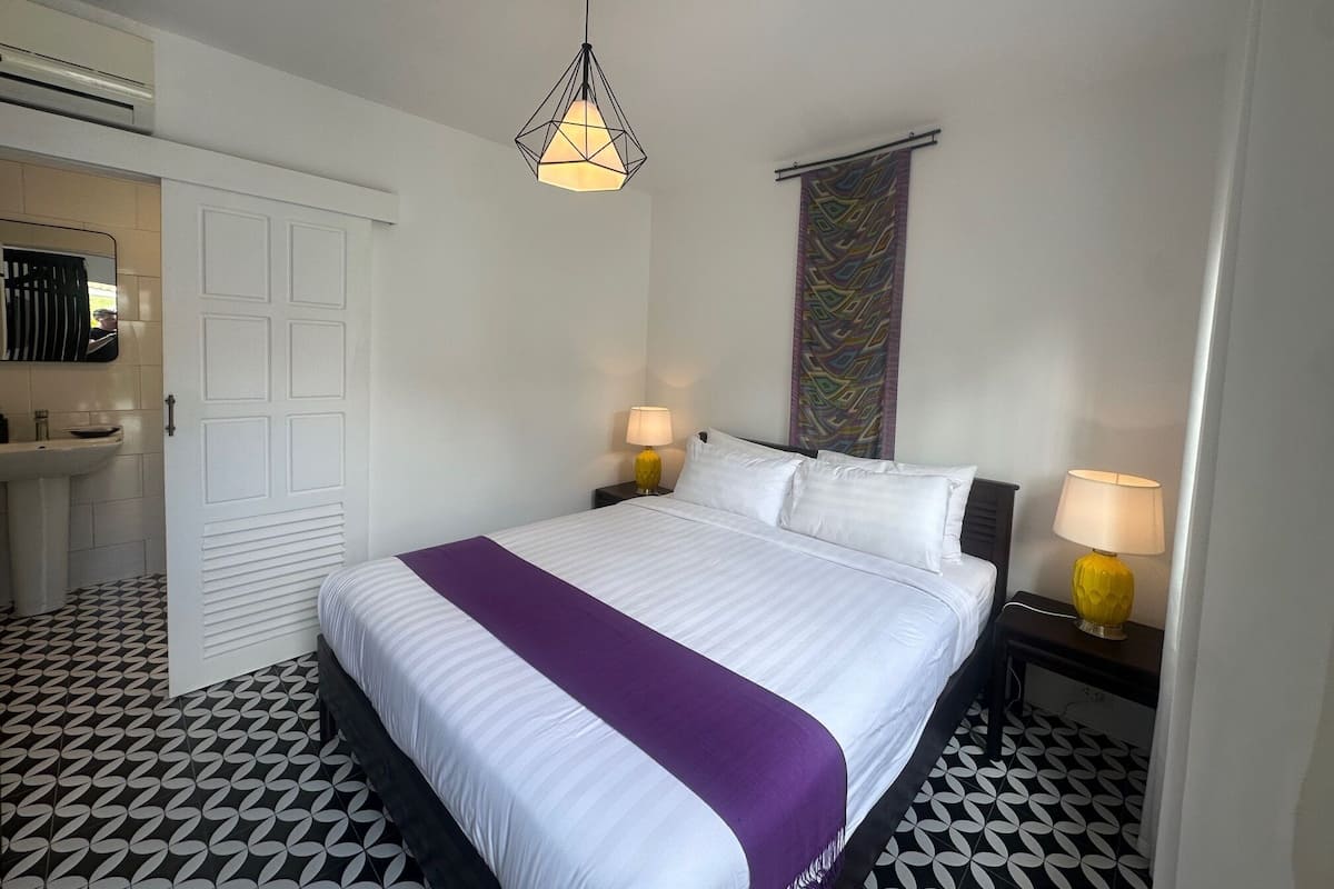 A hotel room with a bed, two bedside lamps, a hanging light fixture, a bathroom with a sink, and a patterned floor.