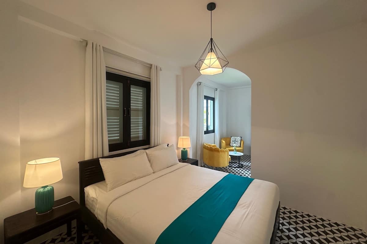A hotel room with a bed, a nightstand, a lamp, a window with blinds, and a hanging light fixture.