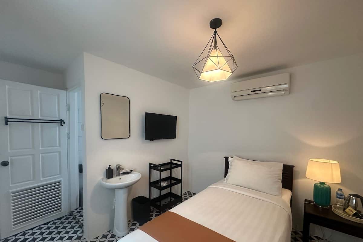 A hotel room with a bed, a bedside table with a lamp, a TV, a mirror, a sink, and an air conditioning unit.