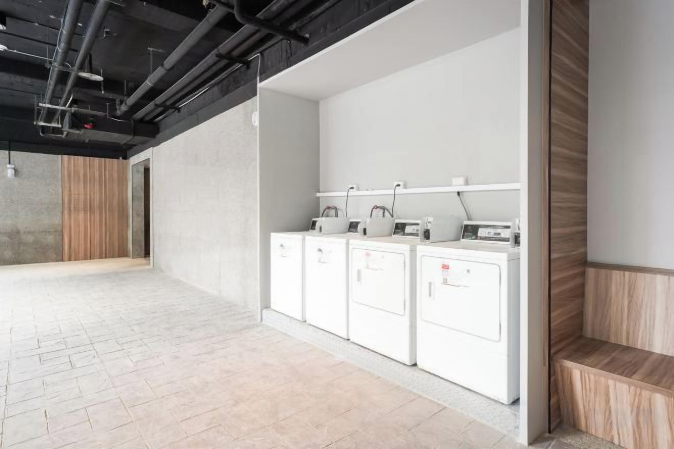 Un área de lavandería con cuatro lavadoras blancas y una secadora, un banco de madera y piso de baldosas.