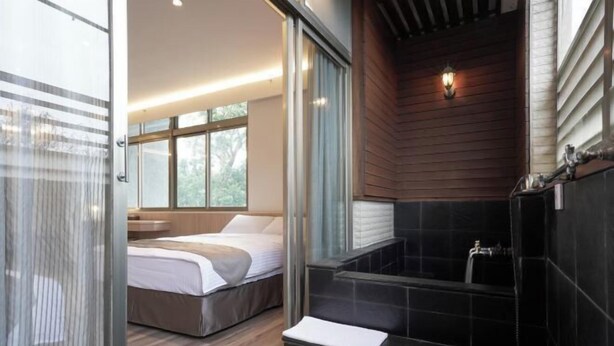 Une salle de bain moderne avec une baignoire, un espace douche et une chambre séparée par une paroi de verre, où se trouve un lit.