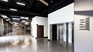 Un couloir moderne et bien éclairé, doté d’un plancher poli, d’un ascenseur en verre et d’un présentoir mural où sont exposés divers objets.