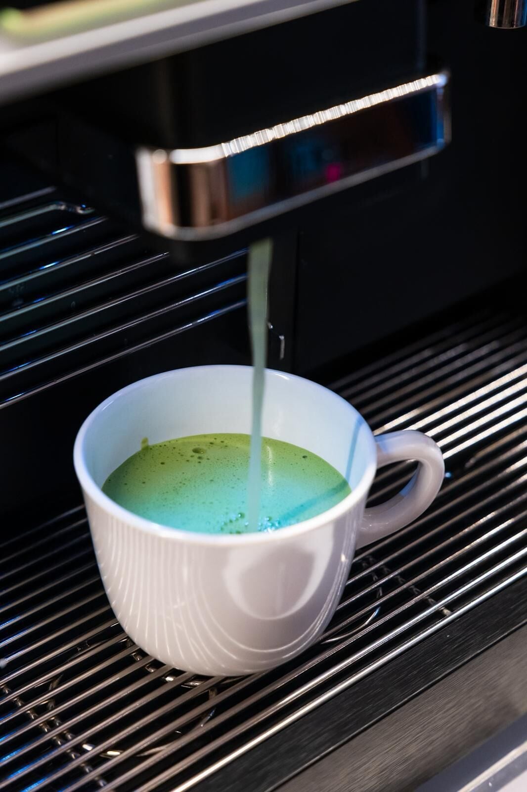 A white cup on a metal rack with a green liquid being dispensed into it.