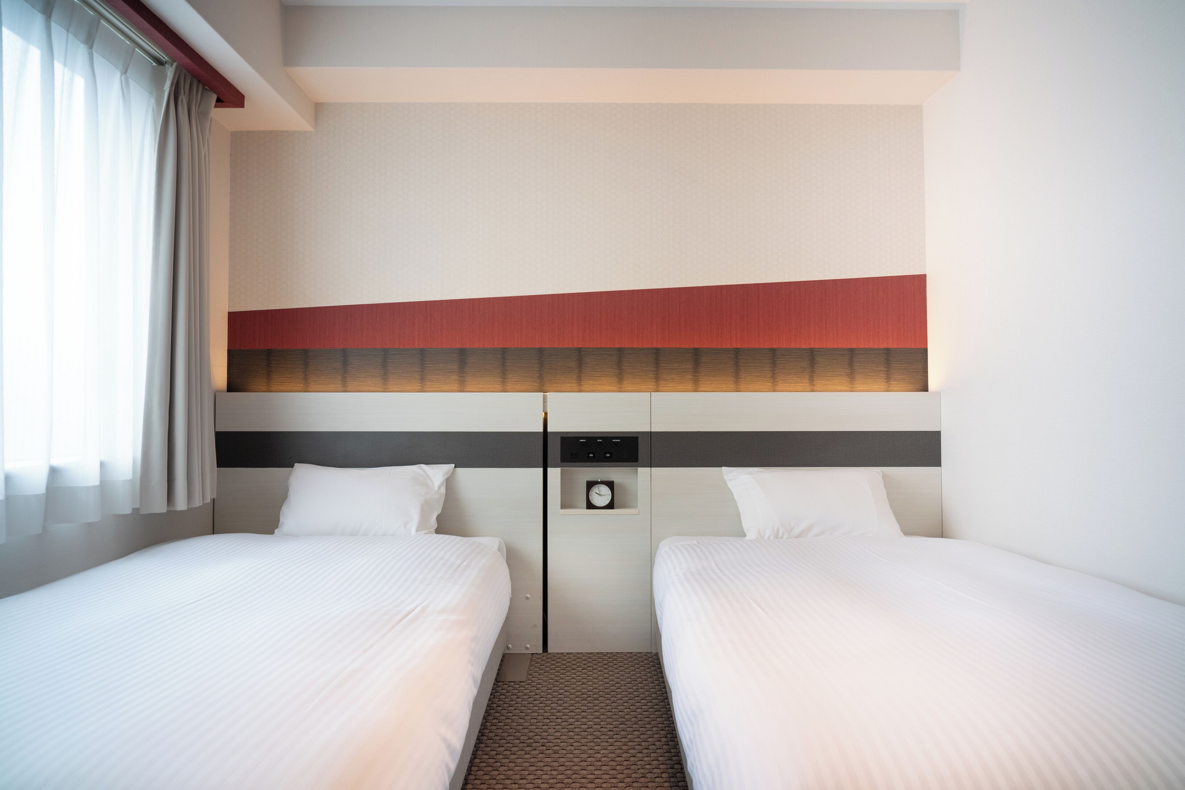 A hotel room with two beds, a headboard with a clock, and a small shelf.
