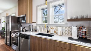 Una cocina moderna con electrodomésticos de acero inoxidable, encimera blanca, armarios de madera y ventana con vistas a árboles.
