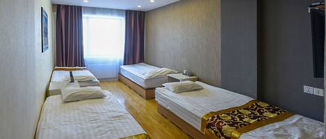 A hotel room with four single beds, a large window with curtains, and a flat-screen TV mounted on the wall.