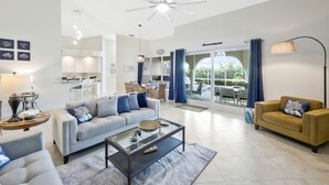 A spacious living room with a modern ceiling fan, a sofa, a coffee table, and a dining area with a view of the outdoors. - Villa Blu- SW Cape Coral- Sleeps 10- Gulf Access Heated Pool Home (Cape Coral)