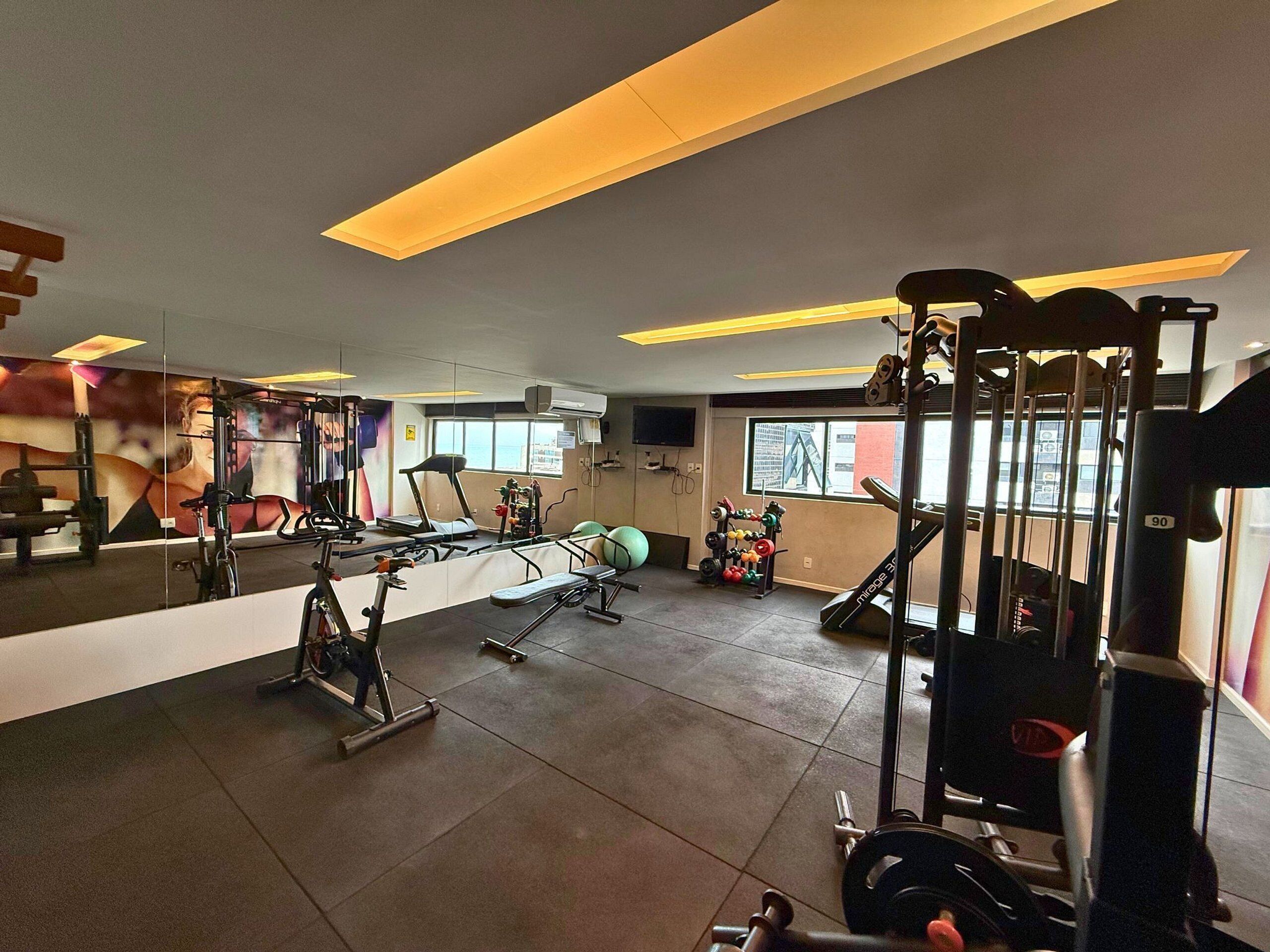 Uma academia com diversos equipamentos de ginástica, um espelho grande e uma janela com vista para o exterior.