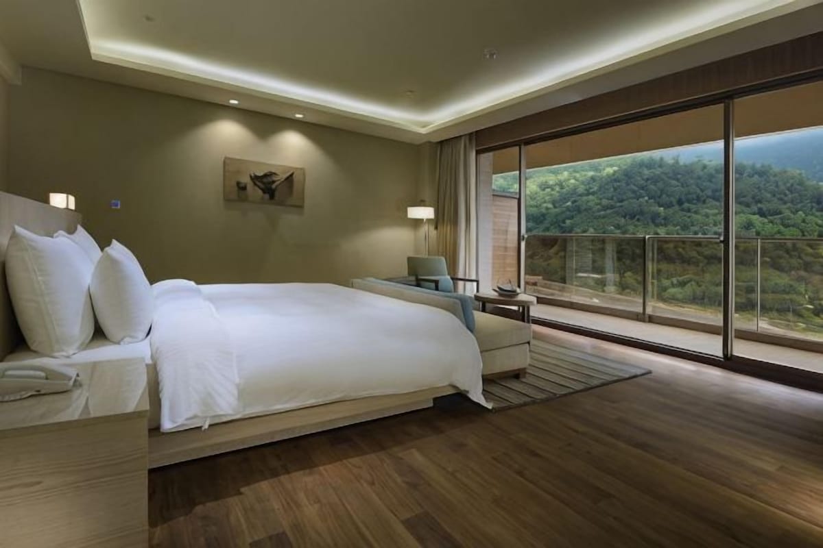 A bedroom with a large bed, a desk, a chair, and a view of a forested area.