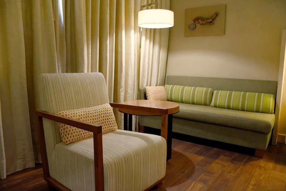 A room with a striped armchair, a wooden side table, a green sofa with striped cushions, and a large window with curtains.