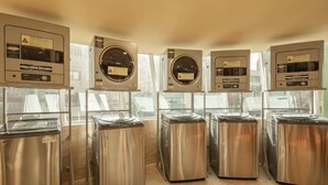 Una fila de lavadoras autoservicio metálicas de acero inoxidable, con paneles de control, ubicada en una sala moderna y bien iluminada con ventanas amplias.