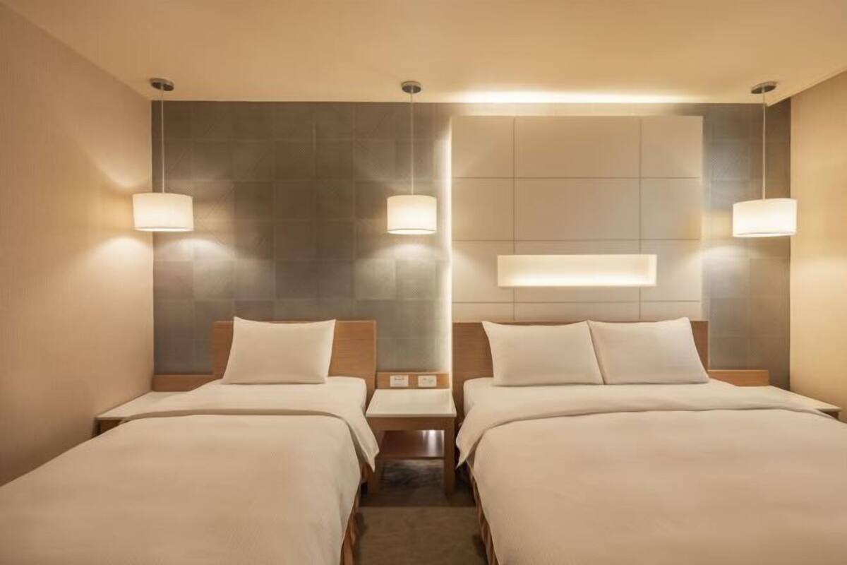A hotel room with two beds, a headboard, bedside tables, and wall-mounted lamps.