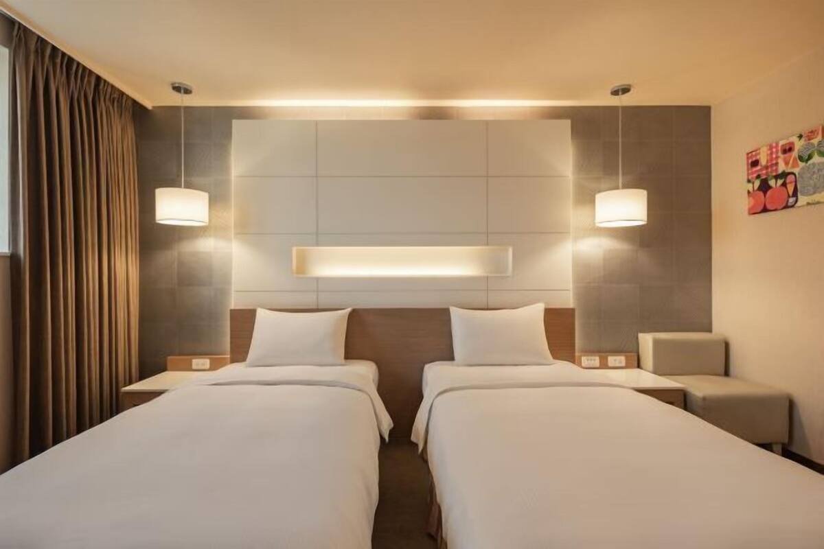 A hotel room with two beds, a headboard with integrated lighting, bedside tables, and a wall-mounted artwork.