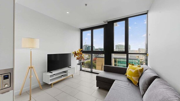 A modern living room with a grey sofa, a TV on a white stand, a floor lamp, and a large window offering a city view. - 1Bed + Car Park Valley Spot (Brisbane)