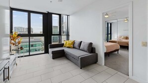 A modern living room with a grey sectional sofa, a glass coffee table, and a view of the city through large windows. - 1Bed + Car Park Valley Spot (Brisbane)