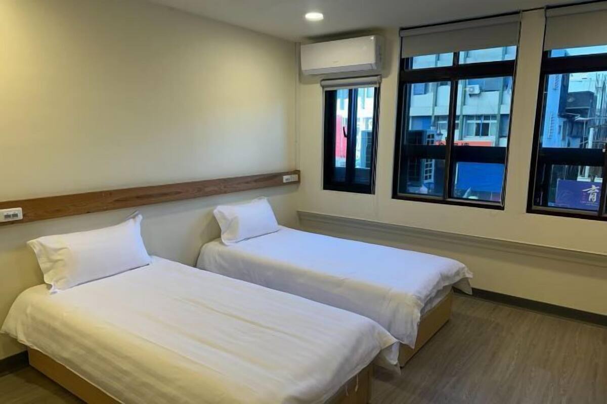 Ein Hotelzimmer mit zwei Einzelbetten, einem Holz-Kopfteil, einer Klimaanlage und Fenstern mit Blick auf Gebäude.