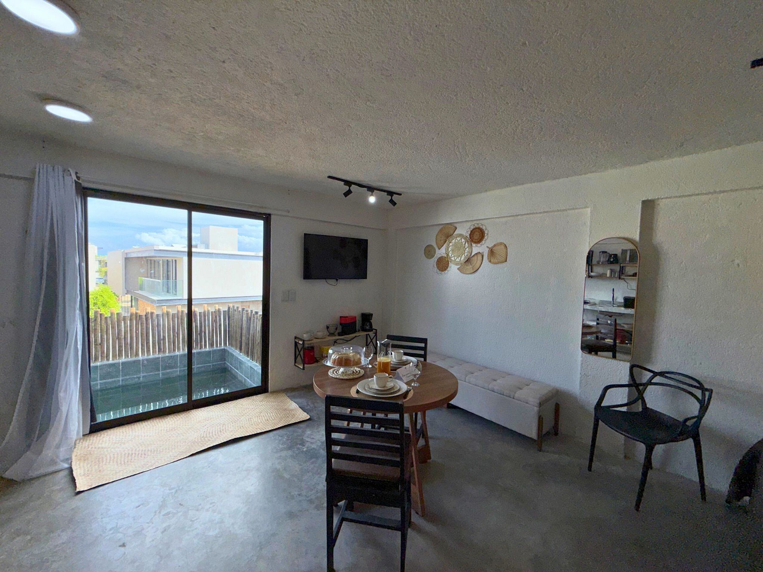 A dining area with a round table, chairs, a bench, a TV, and a view through a sliding door.