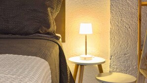 Een netjes opgemaakt bed met hoofdbord, een bedlampje en een klein rond tafeltje met een lamp.