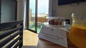 Een kamer met een groot raam, een houten bank, een tafel met een taart en een karaf sinaasappelsap, en een bordje met het kamer nummer en de namen van de gasten.