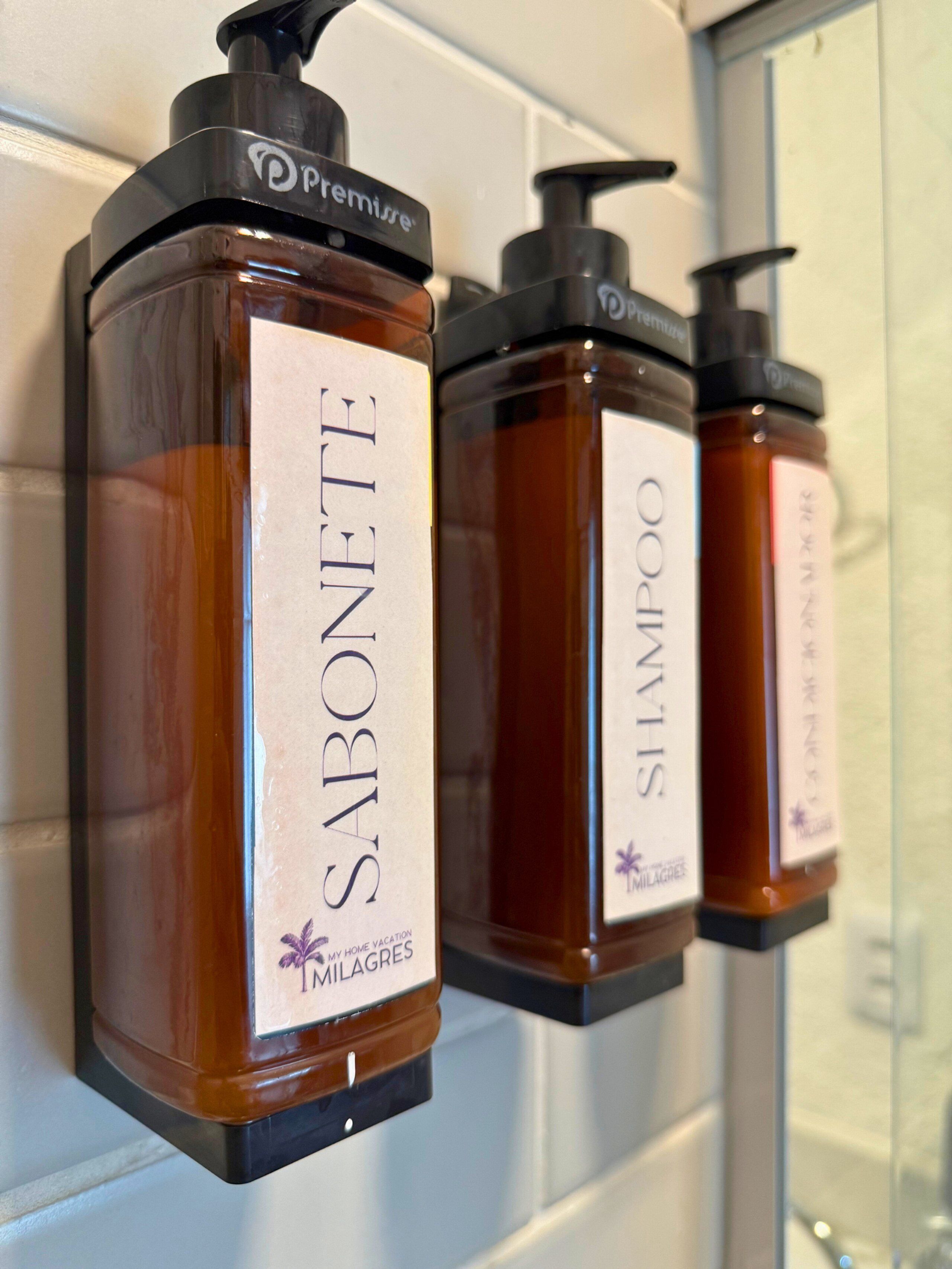 Three bottles of Milagres products: Sabonete, Shampoo, and Shampoos, mounted on a white tiled wall.