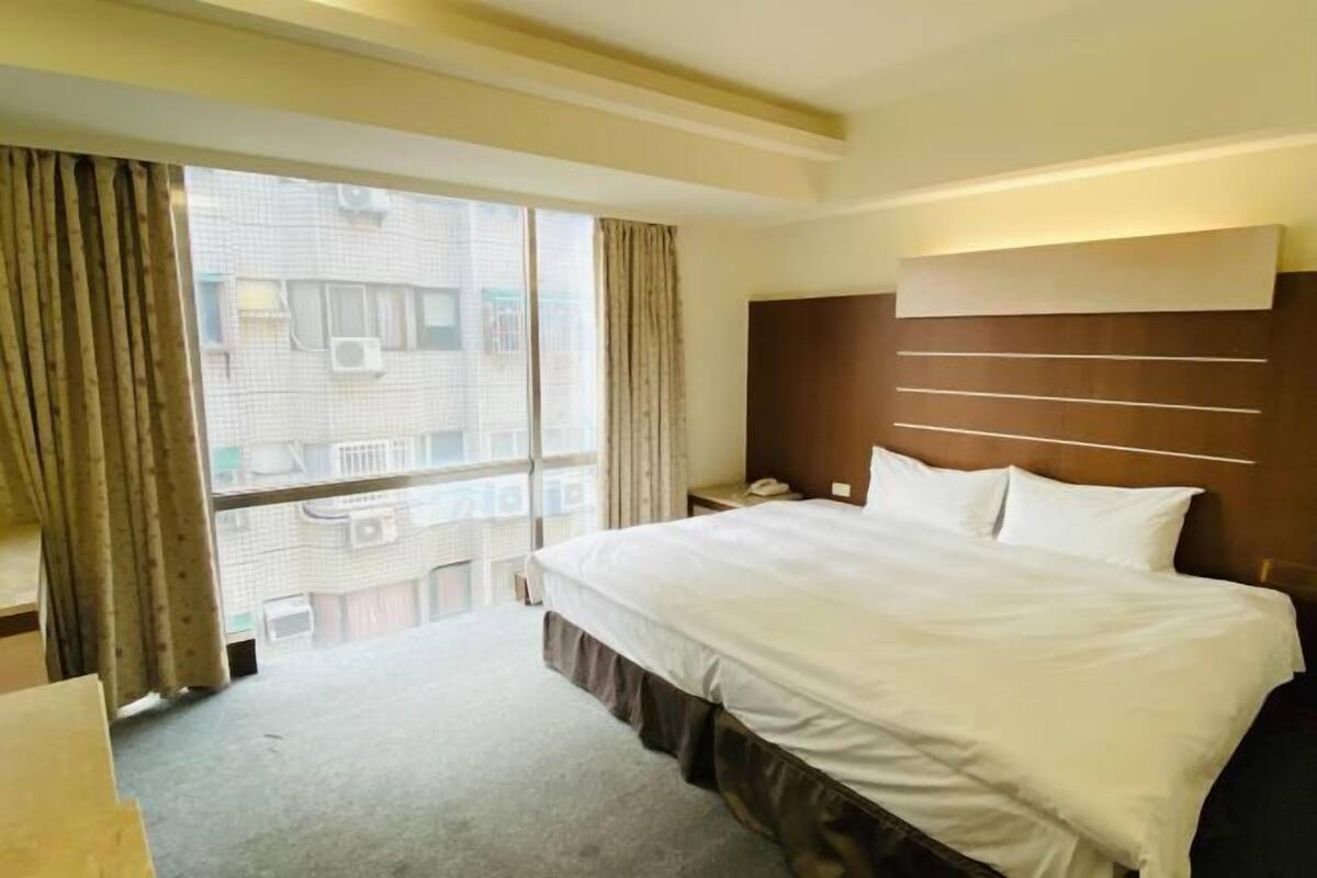 Camera d'albergo con un letto grande, un comodino con una lampada, una finestra con tende e vista su un edificio.