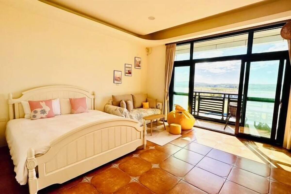 A bedroom with a bed, a bench, a chair, and a large window with a view.