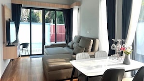 Un salon moderne comprenant un canapé, une table à manger et des chaises. On y trouve une grande fenêtre avec des rideaux, un téléviseur et une petite table ornée d’objets décoratifs.
