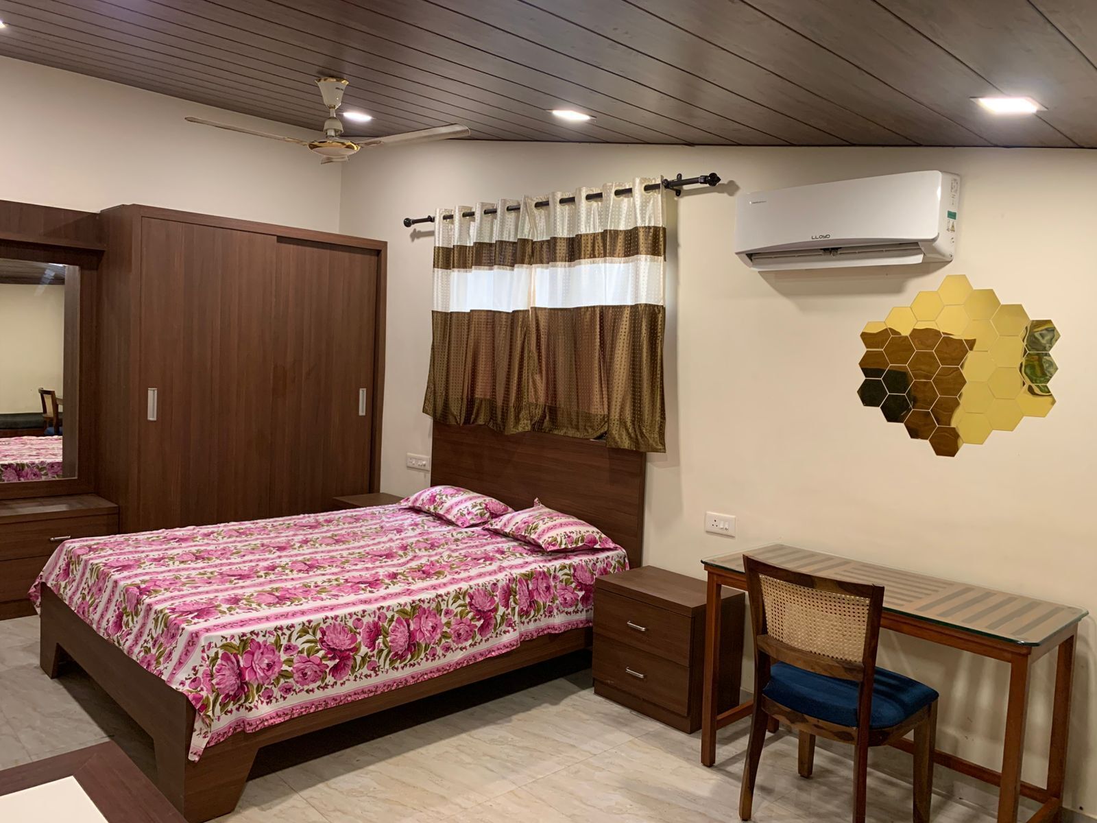Un dormitorio con cama, escritorio, silla, armario, espejo, aire acondicionado y ventilador de techo.