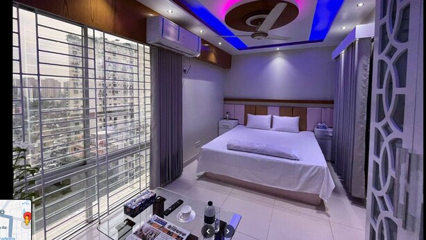Habitació d'hotel moderna amb un llit gran, un ventilador de sostre i vista a la ciutat a través de finestres de sostre a terra.
