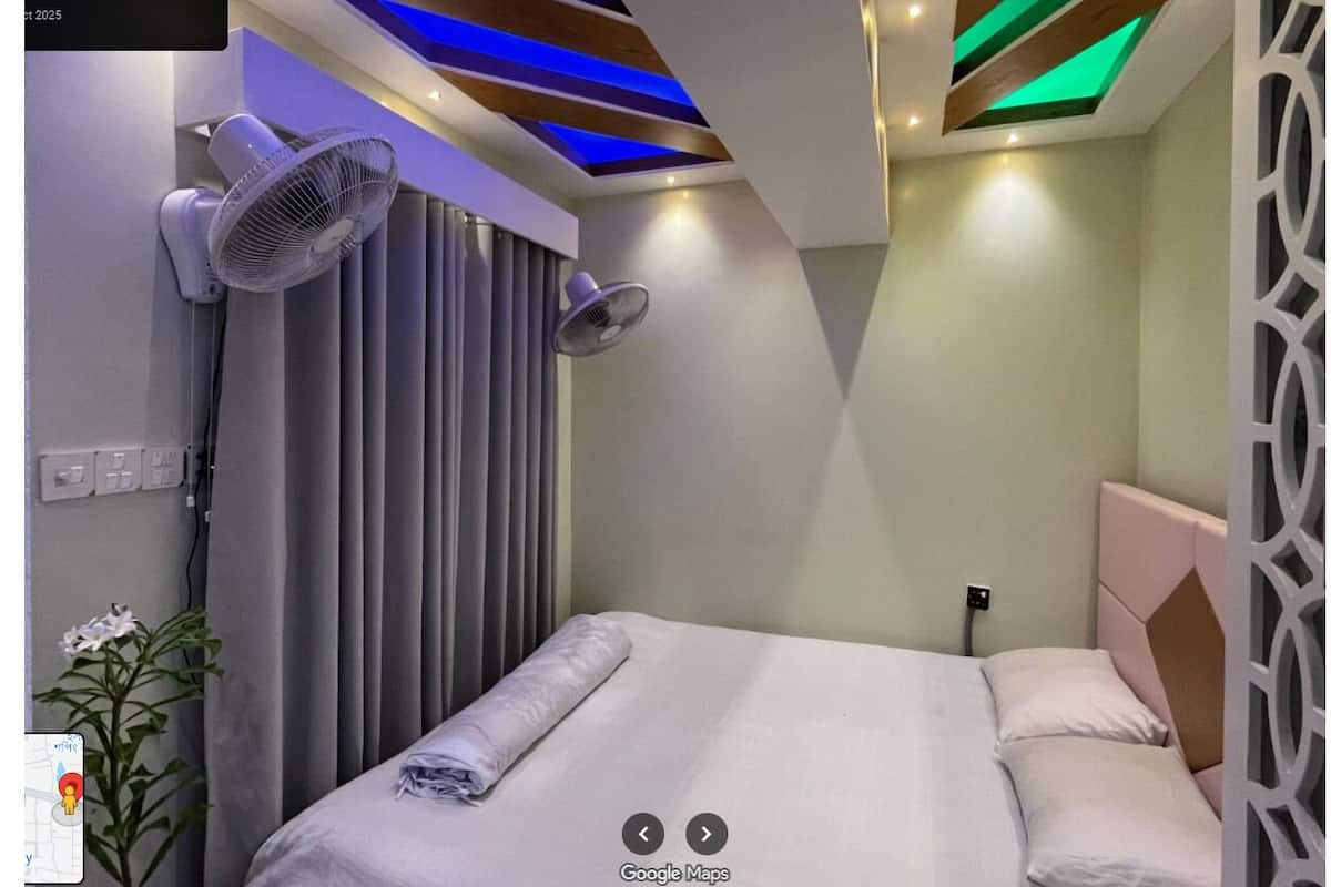 A hotel room with a bed, two wall-mounted fans, a ceiling with blue and green lighting, and a decorative wall panel.