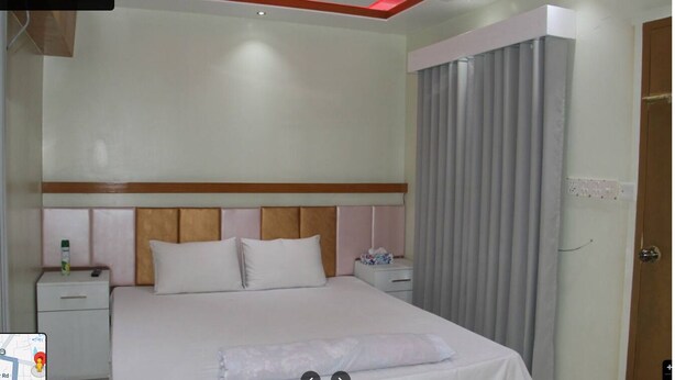 A hotel room with a large bed, a wooden headboard, a nightstand with a bottle, and a curtain. - Hotel The Manha LTD Purana Paltan (Dhaka)