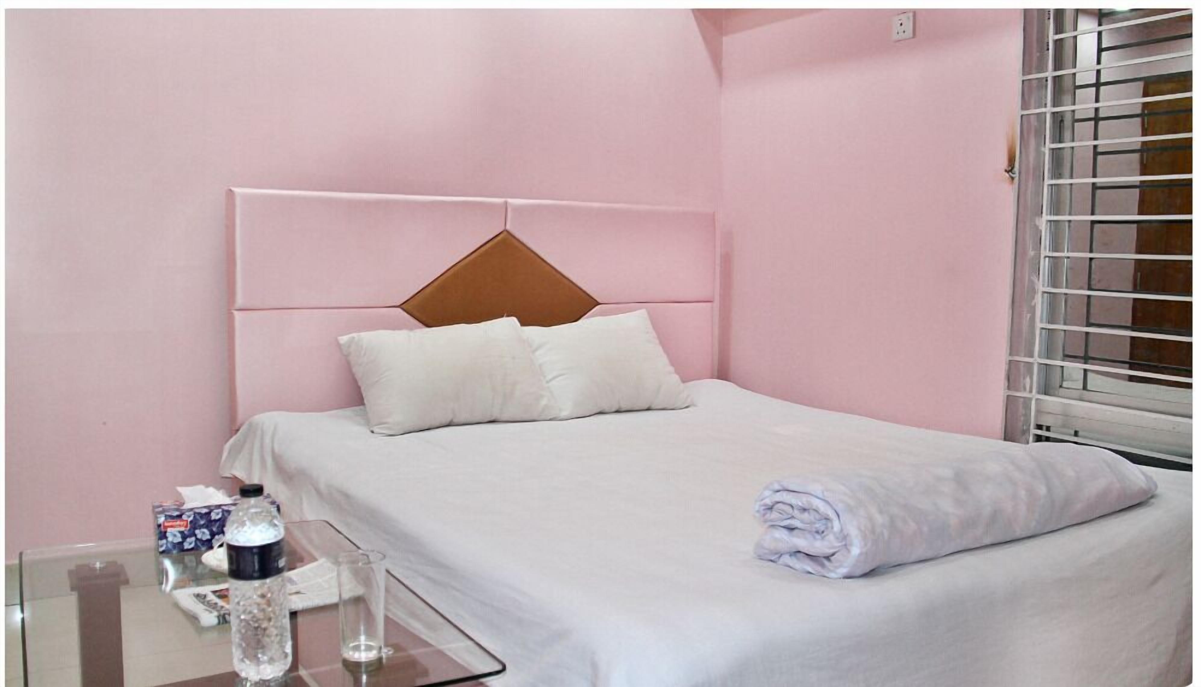 A neatly made bed with white bedding and pillows, a glass table with a bottle and tissue box, and a window with blinds.