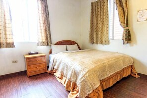 A bedroom with a wooden floor, a bed with a patterned bedspread, a nightstand, and curtains on the windows. - Liching Garden Village (Nantou)