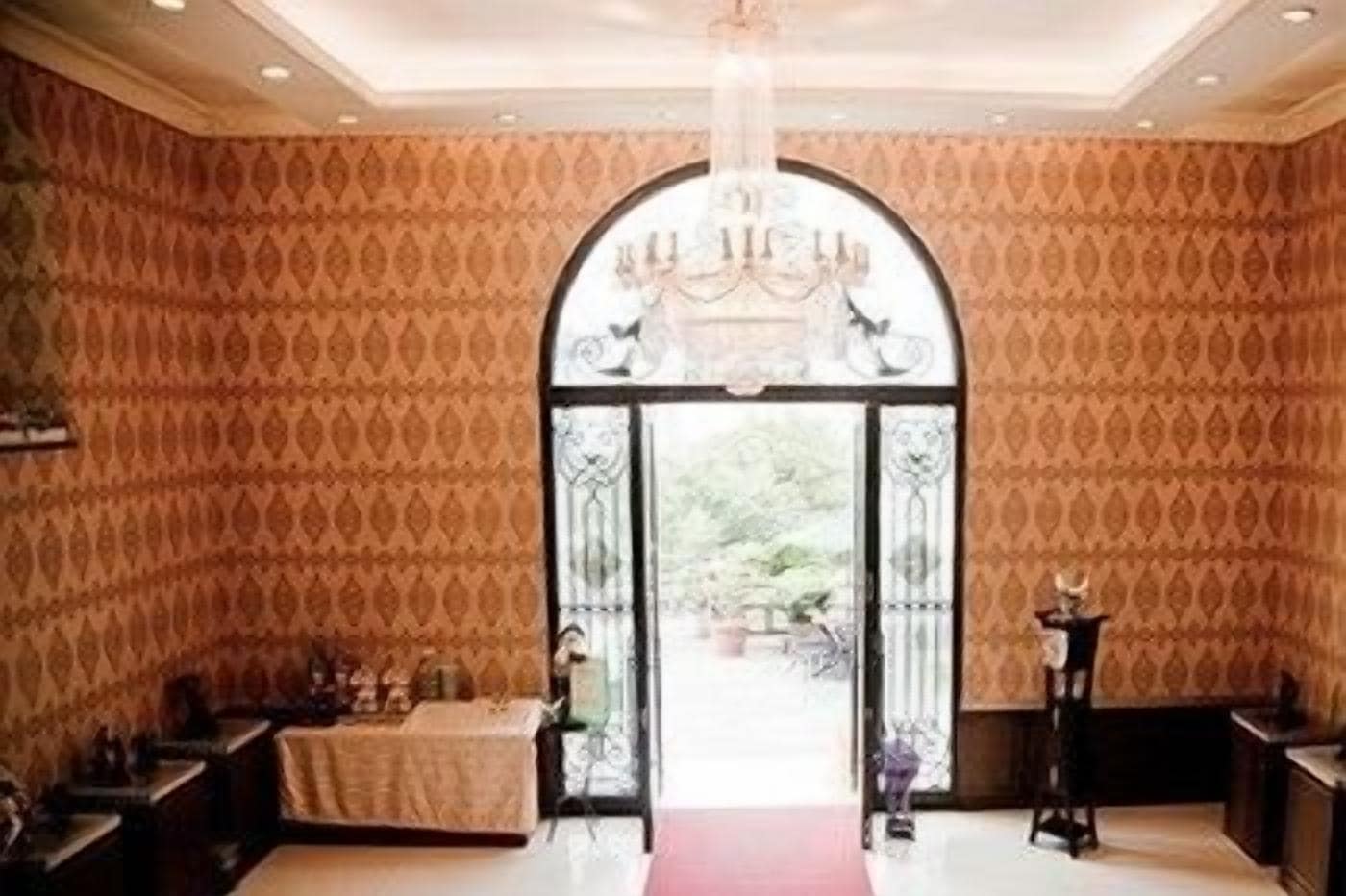 An interior with a patterned wallpaper, a chandelier, and a red carpet leading to an arched doorway.