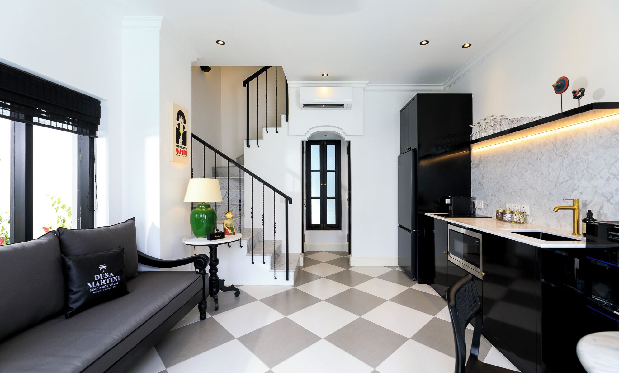 A modern kitchen with a black refrigerator, a marble countertop, and a sink. There is a staircase leading to a second floor.