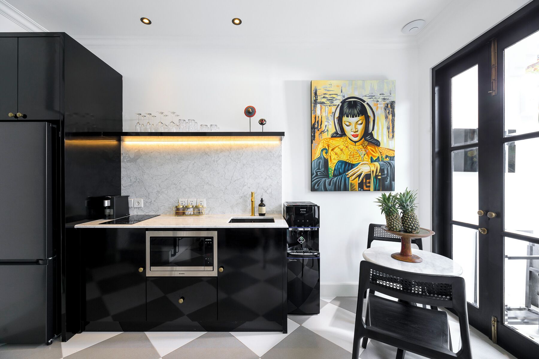 A modern kitchen with black cabinetry, a marble countertop, and a coffee machine. There is a painting on the wall and a window with a view of the outside.